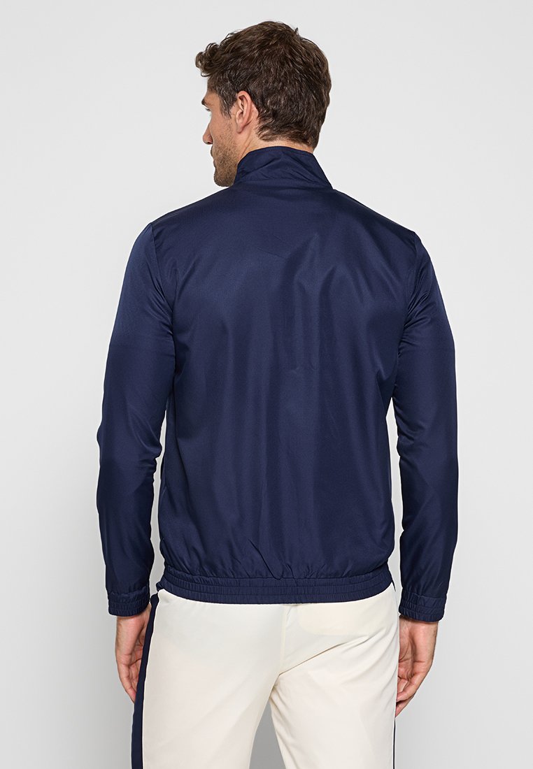 Giacca leggera blu navy con colletto alto, maniche lunghe, polsini elasticizzati e orlo arricciato. Tessuto liscio e lucido senza motivi visibili.