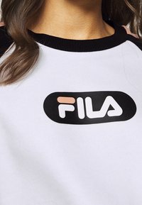 Sweatshirt blanc avec col côtelé noir, arborant un logo noir avec le mot "FILA" en blanc et des accents roses au centre.