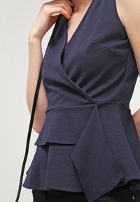 Námořnická modrá peplum blůza s výstřihem do V, která obsahuje kapsu a vrstvené volánkové detaily na lemu, vyrobená z měkké látky.