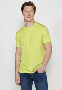 Tricou scurt cu mânecă, de culoare verde lime, dintr-un material moale, cu decolteu rotund și detaliu discret al logo-ului pe piept, asociat cu blugi albaștri.