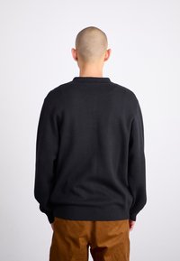 Pull en maille noir avec un col rond, des poignets et un ourlet côtelés, porté sur un pantalon en velours côtelé marron. Manches longues et coupe décontractée.
