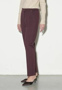Pantaloni tailored bordeaux con una texture liscia, tasche frontali e un design aderente, abbinati a décolleté scamosciate marrone scuro con fiocchi.