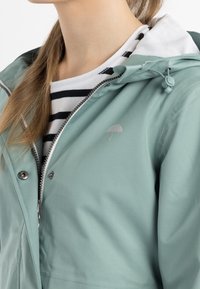 Mintgrön vattentät jacka med dragkedja framtill, silverfärgade detaljer, justerbara snoddar på huvan och en broderad paraplylogotyp.