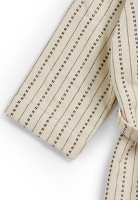 Beige-kangas, jossa on ohuita tummia raidoituksia. Hiha on varustettu ommellulla reunalla ja sidotulla vyöllä, mikä lisää tekstuuria ja yksityiskohtia suunnitteluun.