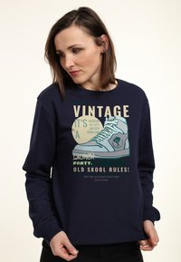 Námořnická modrá mikina s velkým grafickým motivem tenisky a textovými prvky v různých barvách. Dlouhé rukávy a žebrované manžety.