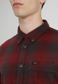 Camicia di flanella a quadri rossi e neri con colletto button-down, due tasche frontali e bottoni neri, con un piccolo logo Lee sulla tasca del petto.