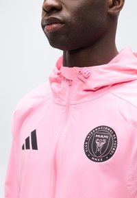 Κοντινό πλάνο ενός ατόμου που φοράει ροζ μπουφάν με φερμουάρ και κουκούλα, με τα λογότυπα του Club Internacional de Fútbol Miami και της Adidas στο στήθος.