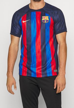 Mann trägt ein FC Barcelona Fußballtrikot mit kurzen Ärmeln, roten und blauen vertikalen Streifen und schwarzen Hosen, steht vor einem einfarbigen Hintergrund.