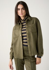 Olive green suede-like shirt with two front pockets, snap closures, and a relaxed fit, layered over a navy and olive striped turtleneck.