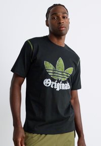 Férfi fekete pólóban, amelyen elkopott zöld Adidas trefoil logó és "Originals" felirat látható, olíva zöld nadrággal párosítva.