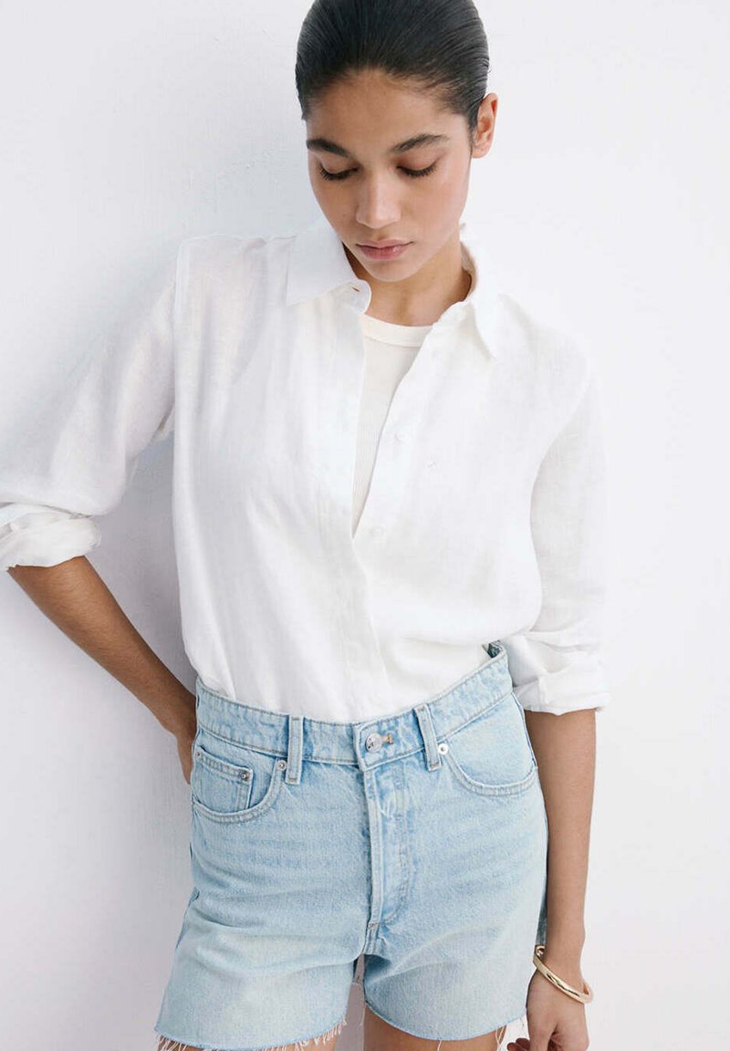 Femme aux cheveux foncés portant une chemise blanche boutonnée et un short en jean bleu clair, debout contre un mur blanc, regardant vers le bas avec une main sur la hanche.