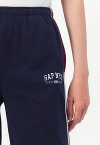 Närbild av en person som bär marinblå sweatpants med vita och vinröda sidostripes och "GAP NYC EST. 1969"-logotyp på låret.