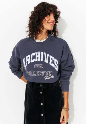 Sweatshirt bleu marine foncé avec l'inscription "COLLECTION ARCHIVES 1975" imprimée en blanc usé, accompagné d'une jupe en velours côtelé noire avec des boutons sur le devant.