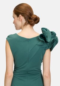 Robe verte avec un bustier ajusté et un grand volant en couches sur une épaule, ainsi qu'une fermeture éclair dissimulée dans le dos. Texture de tissu lisse.