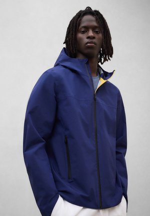 Persona joven con rastas que lleva una chaqueta azul con capucha y cremallera, con forro amarillo, sobre una camiseta gris, de pie frente a un fondo gris liso.