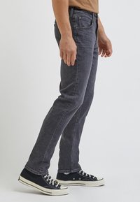 Jeans de mezclilla gris con un diseño ajustado, que cuentan con bolsillos laterales y detalles de costura sutiles, combinados con zapatillas de lona negras.