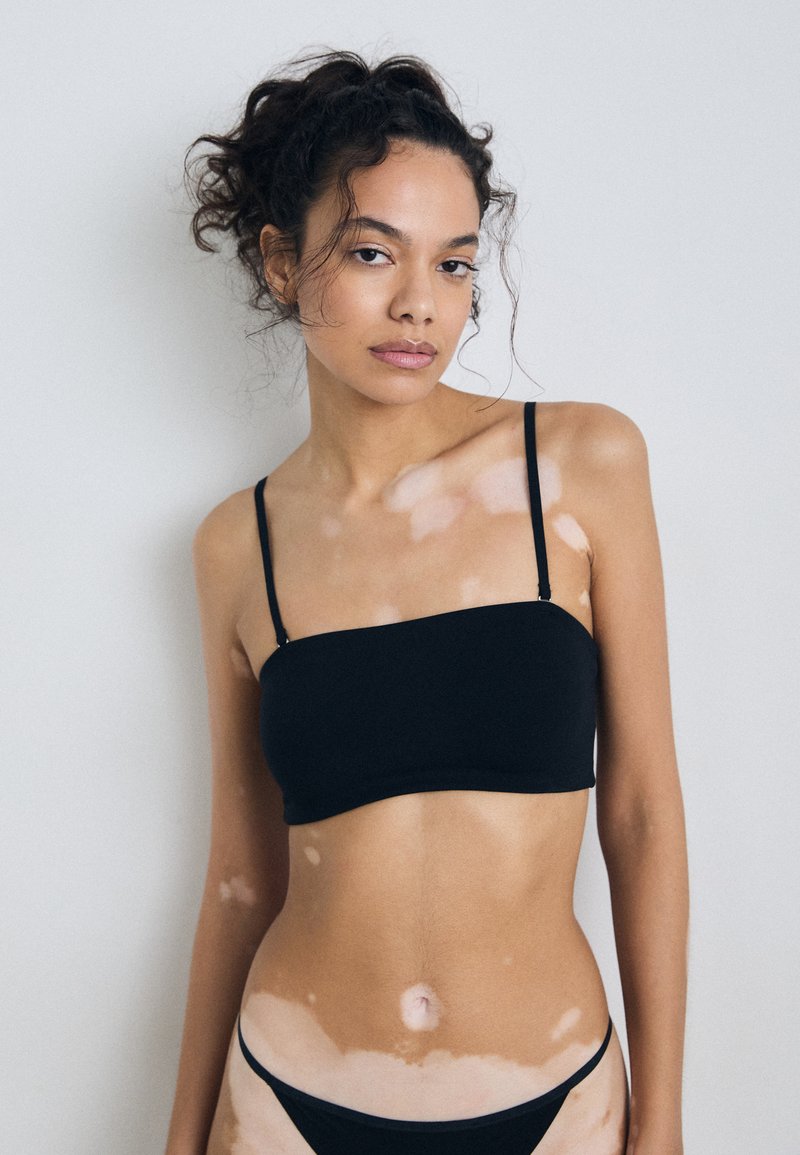 Bralette noir sans couture avec des fines bretelles réglables, un décolleté droit et une texture lisse. Porté avec un bas de bikini noir assorti.