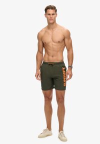 Durchtrainierter, oberkörperfreier Mann in olivgrünen Shorts mit orangefarbener "SUPERDRY"-Aufschrift, beigen Turnschuhen und Armbanduhr, stehend vor weißem Hintergrund.