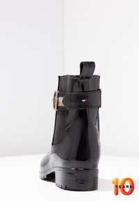 Botín negro brillante hasta el tobillo con paneles elásticos laterales, hebilla decorativa y suela texturizada. Presenta una lengüeta en el talón.