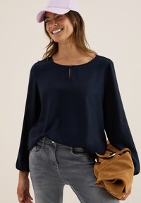 Blusa de satén azul marino de manga larga con escote en forma de llave. Combinada con unos jeans de mezclilla gris y un bolso de ante color beige, complementada con una gorra lila clara.