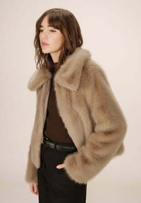 Giacca in pelliccia ecologica color marrone chiaro con un ampio colletto, dotata di zip e un tessuto a coste sotto, abbinata a pantaloni neri e una cintura.