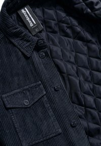 Giacca camicia in corduroy blu marino con rivestimento trapuntato. Presenta un colletto, chiusura con bottoni e una tasca sul petto con pattina. Tessuto testurizzato.