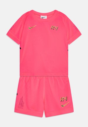 Růžová sportovní souprava Nike, která obsahuje tričko s krátkým rukávem a kraťasy. Zlaté loga a akcenty. Hladká textura s grafickým motivem plamene na kraťasech.