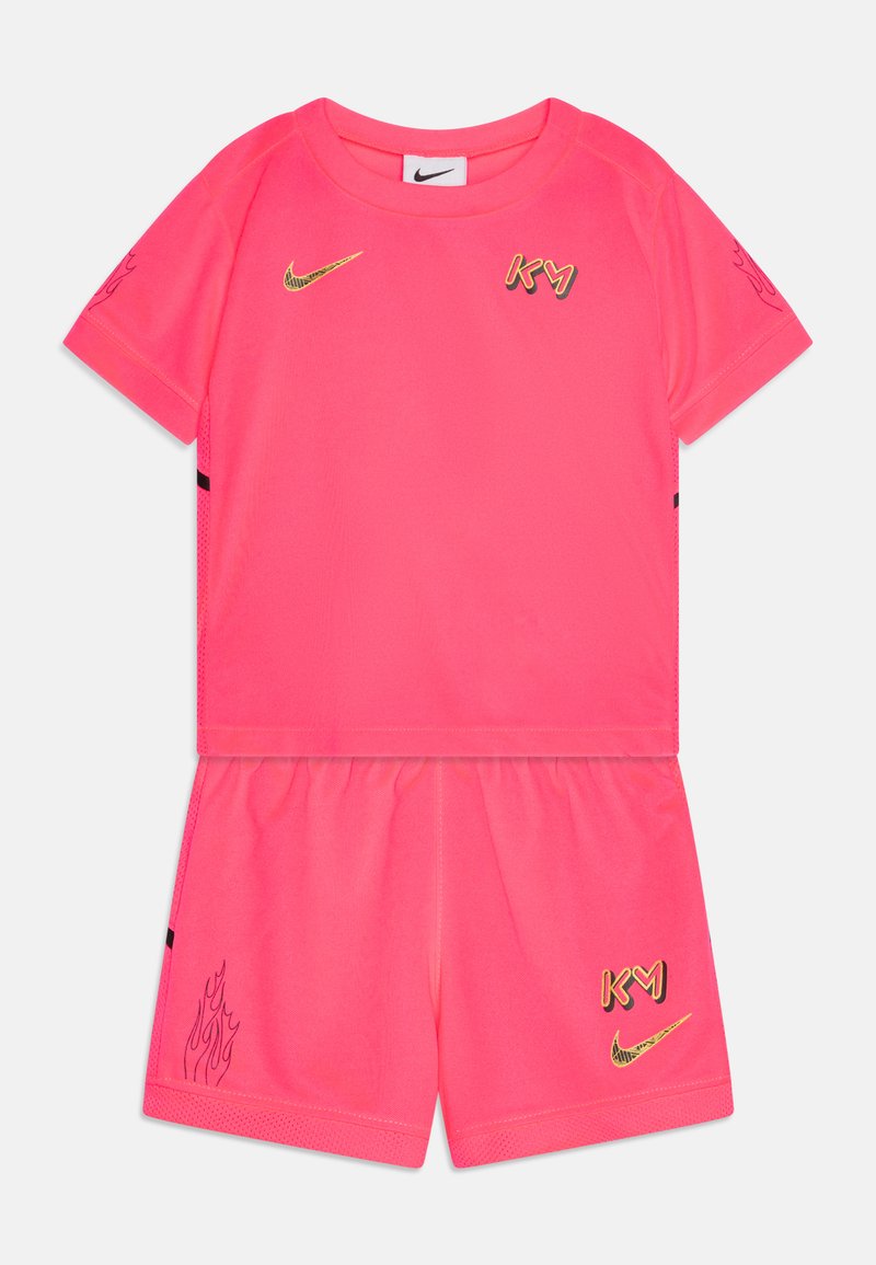 Roze Nike sporta komplekts, kas sastāv no īsām piedurknēm, topa un šortiem. Zelta logotipi un akcenti. Gluda tekstūra ar liesmu grafiku šortos.