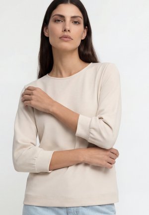 Kvinde med langt mørkt hår iført en lysebeige langærmet skjorte og lyseblå jeans, poserende med armene krydsede mod hvid baggrund.