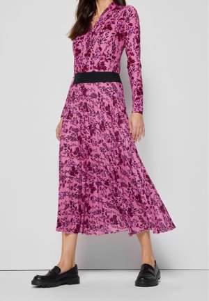 Langwerpige, roze bloemenjurk met lange mouwen en een zwarte tailleband, met een geplisseerde rok en een gladde, lichte stof. Zwarte instappers maken de look af.