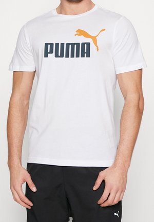 Hombre vistiendo una camiseta blanca de Puma con el logo naranja y pantalones cortos negros de Puma frente a un fondo liso.