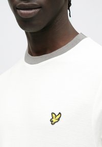 Bela srajca s kratkimi rokavi, sivim rebrastim ovratnikom in majhno črno-rumeno vezeno ptičjo značko na levi strani prsi. Gladek tekstura.