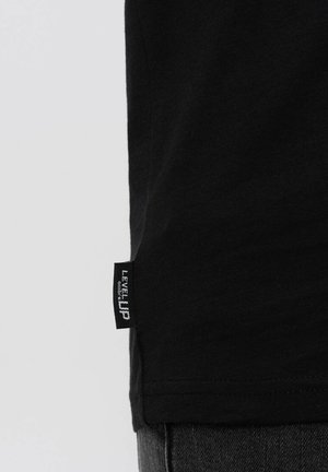Camiseta de algodón negra con cuello redondo, etiqueta en la costura lateral que dice "LEVEL UP" y una textura suave.