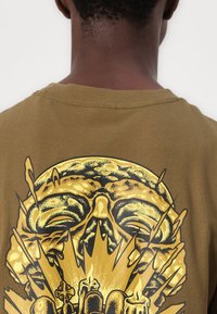 Olivenfarget bomulls T-skjorte med et stort gull- og svart grafisk design av et hode, pigger og lys på baksiden.