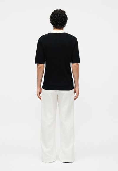 Άνδρας με σγουρά μαλλιά που φοράει μαύρο κοντομάνικο πουκάμισο και λευκό παντελόνι με φαρδιά πόδια, στέκεται με την πλάτη προς τα εμπρός σε απλό φόντο.