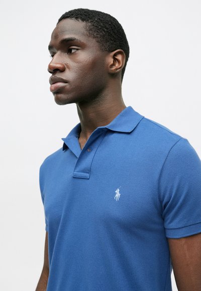 Μπλε πόλο μπλούζα με υφασμάτινη επιφάνεια, κοντά μανίκια και τρεις κουμπάκια. Διαθέτει ένα μικρό λευκό λογότυπο στο στήθος.