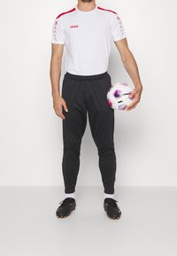 Man som håller en vit och lila Nike fotboll, klädd i en vit JAKO sporttröja, svarta byxor, vita strumpor och svarta fotbollsskor.