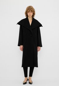 Lungo cappotto nero con ampio colletto, chiusura a un bottone e tasche laterali. Indossato sopra pantaloni neri aderenti e scarpe nere a punta.