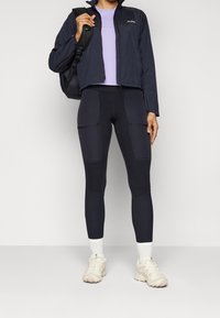 Marinblå lätt jacka över en lilamint topp, i kombination med svarta högmidjade leggings med dragkedjefickor, samt krämfärgade sneakers med vita strumpor.