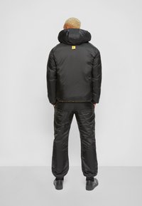 Svart isolerad jacka med en stor huva, prydd med en gul logotyp, i kombination med matchande svarta byxor, båda med en slät yta.