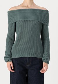 Maglione a spalle scoperte in verde attenuato, texture a coste, maniche lunghe e scollo ampio. Abbinato a pantaloni in denim scuro.