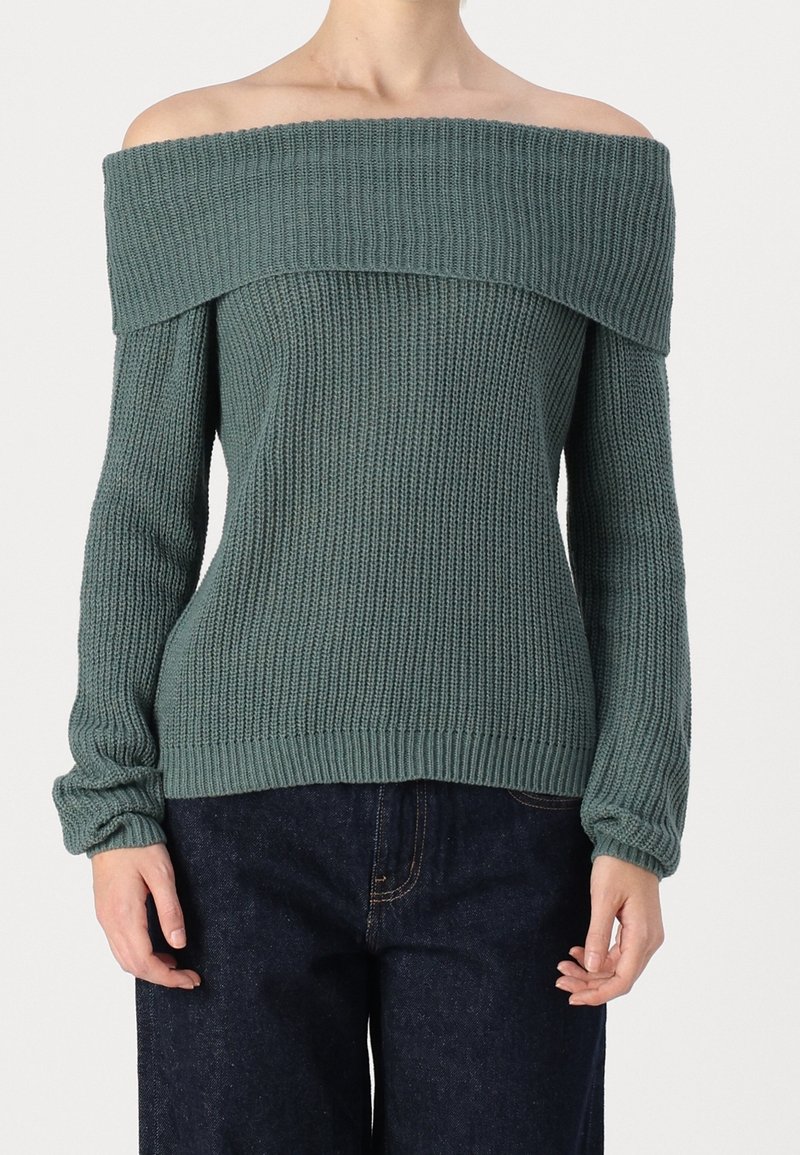 Maglione a spalle scoperte in verde attenuato, texture a coste, maniche lunghe e scollo ampio. Abbinato a pantaloni in denim scuro.