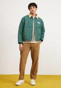Green quilted jacket with beige collar, front button closure, and logo patch. Brown pants and cream shoes with textured soles.