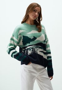 Pull en tricot présentant un design de paysage montagneux dans des tonalités de vert, bleu marine et crème. Coupe ample avec de larges rayures sur les manches et un col côtelé.