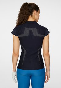 Mörkblå pikétröja med kortärmar, ribbad struktur och meshdetalj på ryggen, bärs med blå byxor och en vit handske.