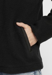 Czarna polarowa kurtka o miękkiej, teksturowanej powierzchni; ma boczną kieszeń z eleganckim, czarnym wykończeniem. Zbliżenie na tkaninę i dłoń.