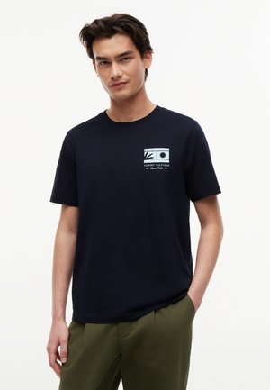 Junger Mann trägt ein schwarzes Tommy Hilfiger T-Shirt mit einer kleinen Grafik von Sonne und Vogel auf der Brust, kombiniert mit olivgrünen Hosen.