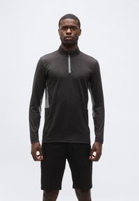 Άνδρας όρθιος, στραμμένος προς τα εμπρός, φοράει μαύρο μακρυμάνικο ζακέτα με φερμουάρ και γκρι πλαϊνές φάσες, καθώς και μαύρο σορτς σε λευκό φόντο.