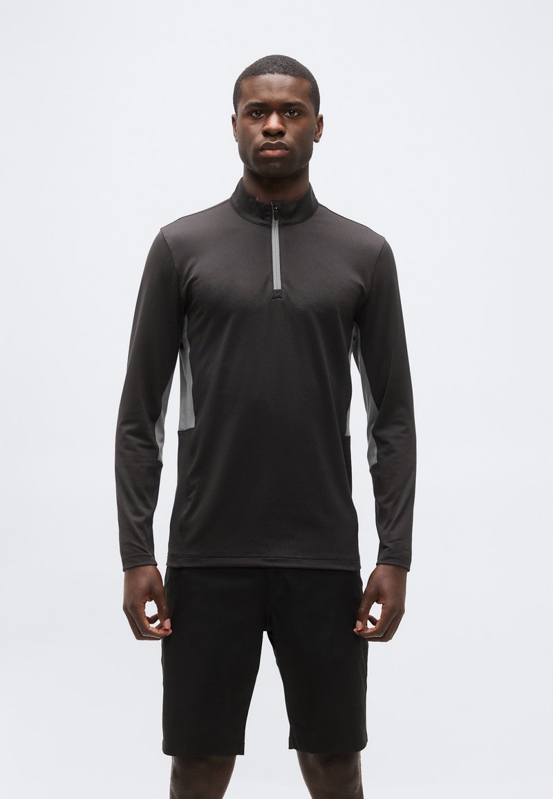 Άνδρας όρθιος, στραμμένος προς τα εμπρός, φοράει μαύρο μακρυμάνικο ζακέτα με φερμουάρ και γκρι πλαϊνές φάσες, καθώς και μαύρο σορτς σε λευκό φόντο.