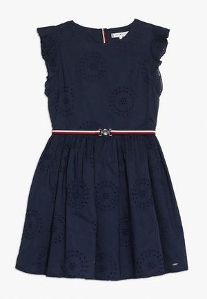 Marineblauwe jurk met een aansluitend lijfje en een uitlopende rok. Voorzien van oogjespatronen, ruches aan de mouwen en een rood-wit gestreepte ceintuur met zilverkleurige details.